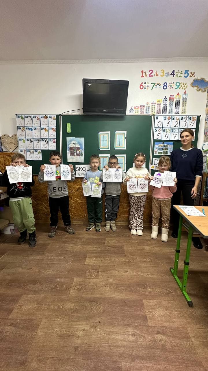 Young children holding up their artwork with their SKOLKA teacher in a decorated kindergarten classroom